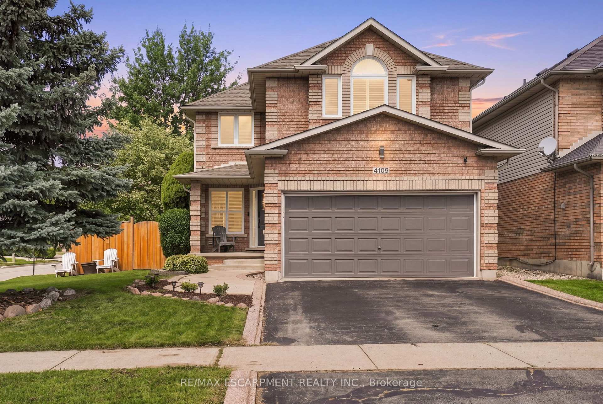 Two-storey brick house with a double garage, landscaped lawn, and mature trees at dusk.