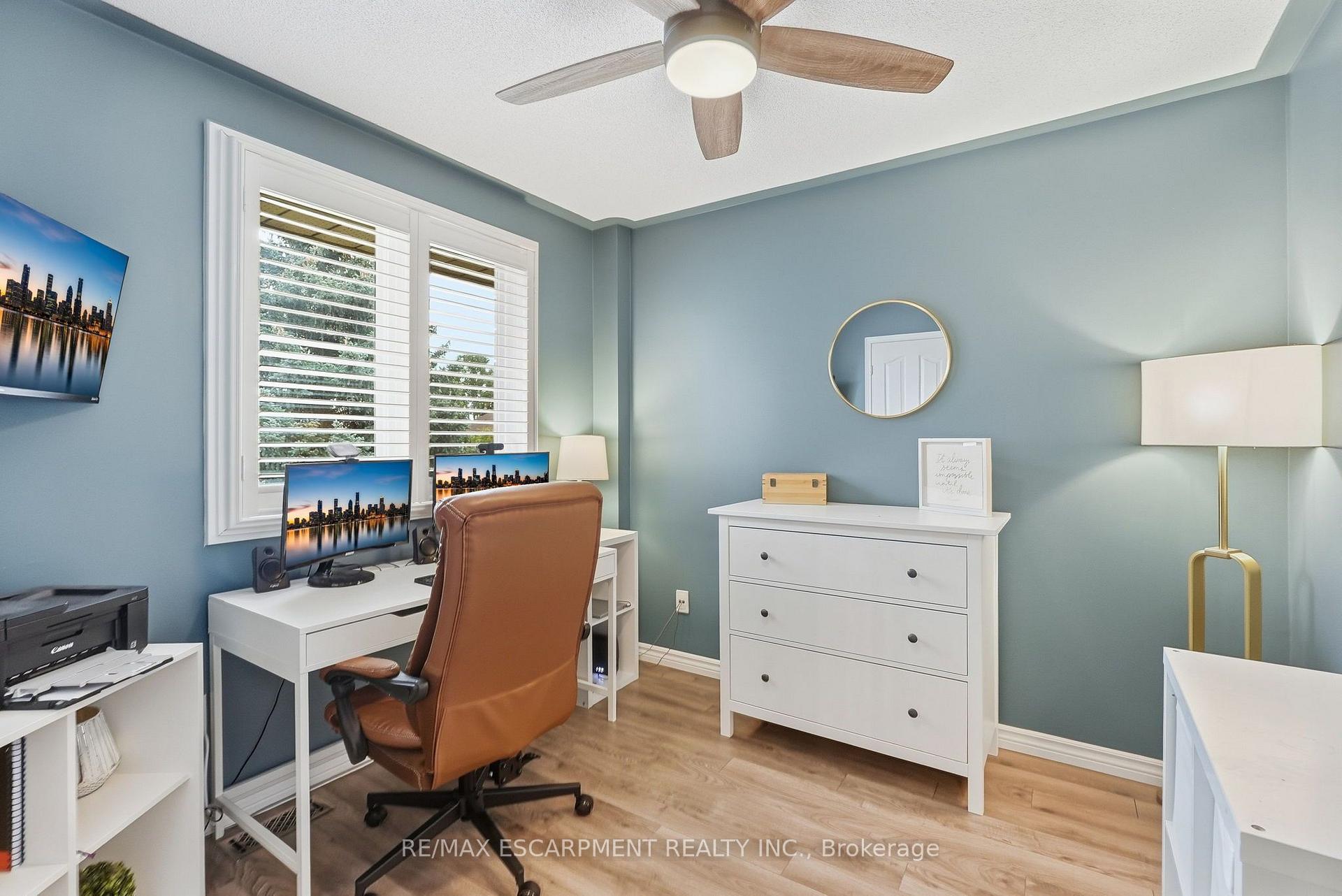 Furnished home office with blue-gray walls, white desk and dresser, brown office chair, and shuttered windows.
