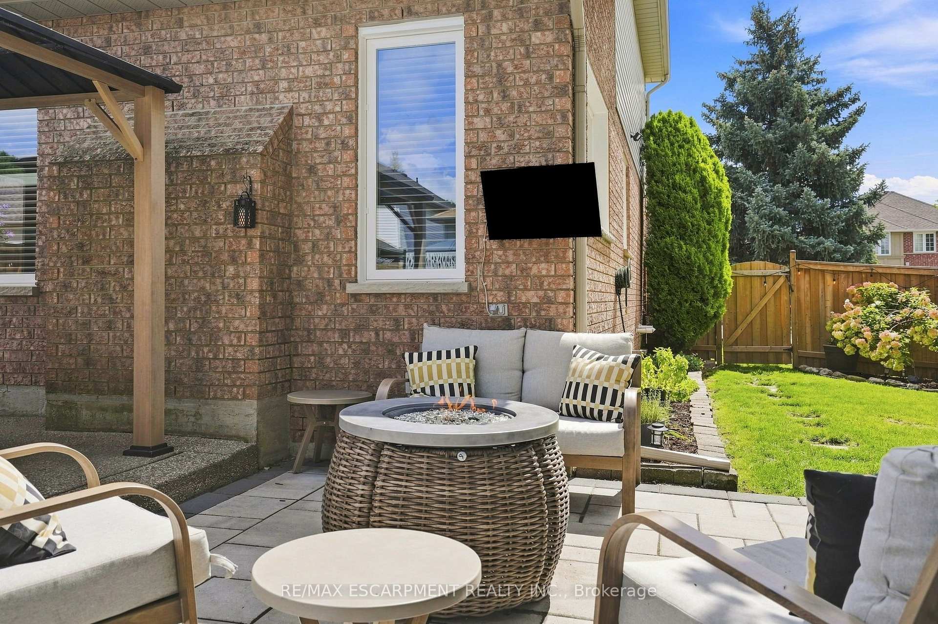 Backyard patio with cushioned seating, a round fire table, and a fenced lawn beside a brick house.