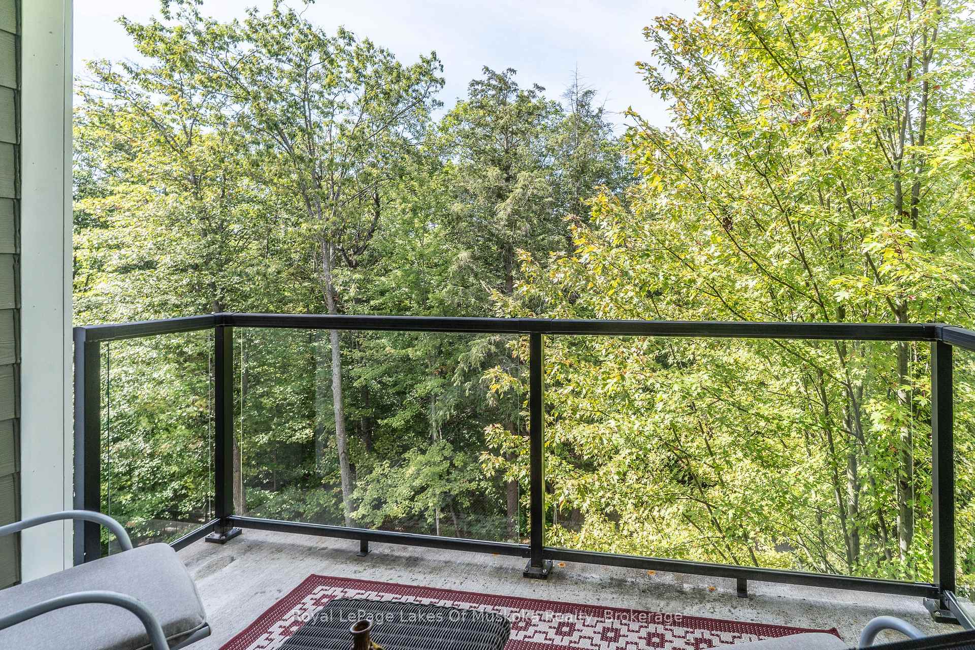 Apartment balcony with glass railing and seating overlooking leafy trees.
