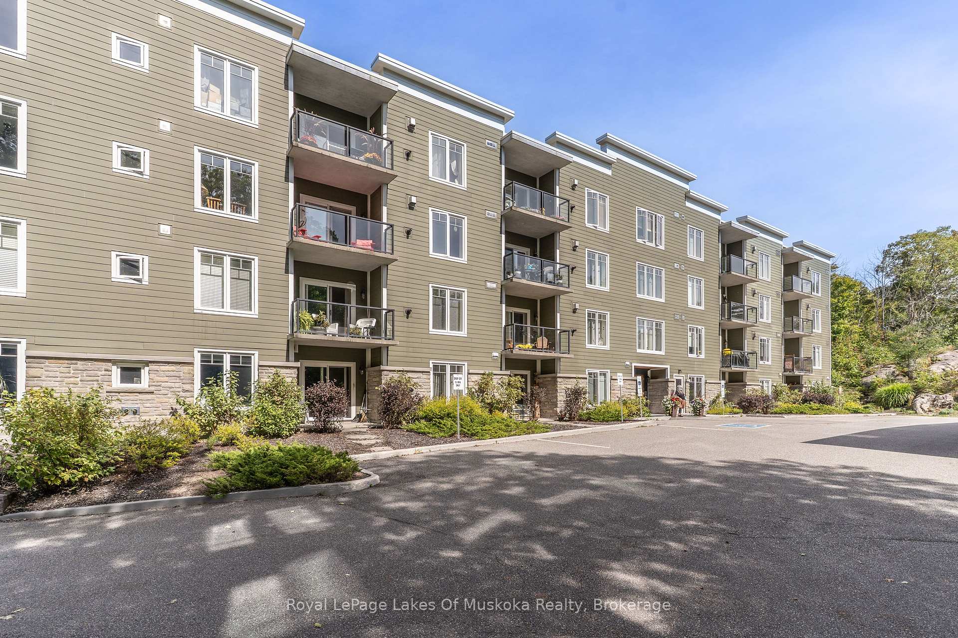 Olive-green apartment building with glass balconies, stone base, and landscaped parking area.