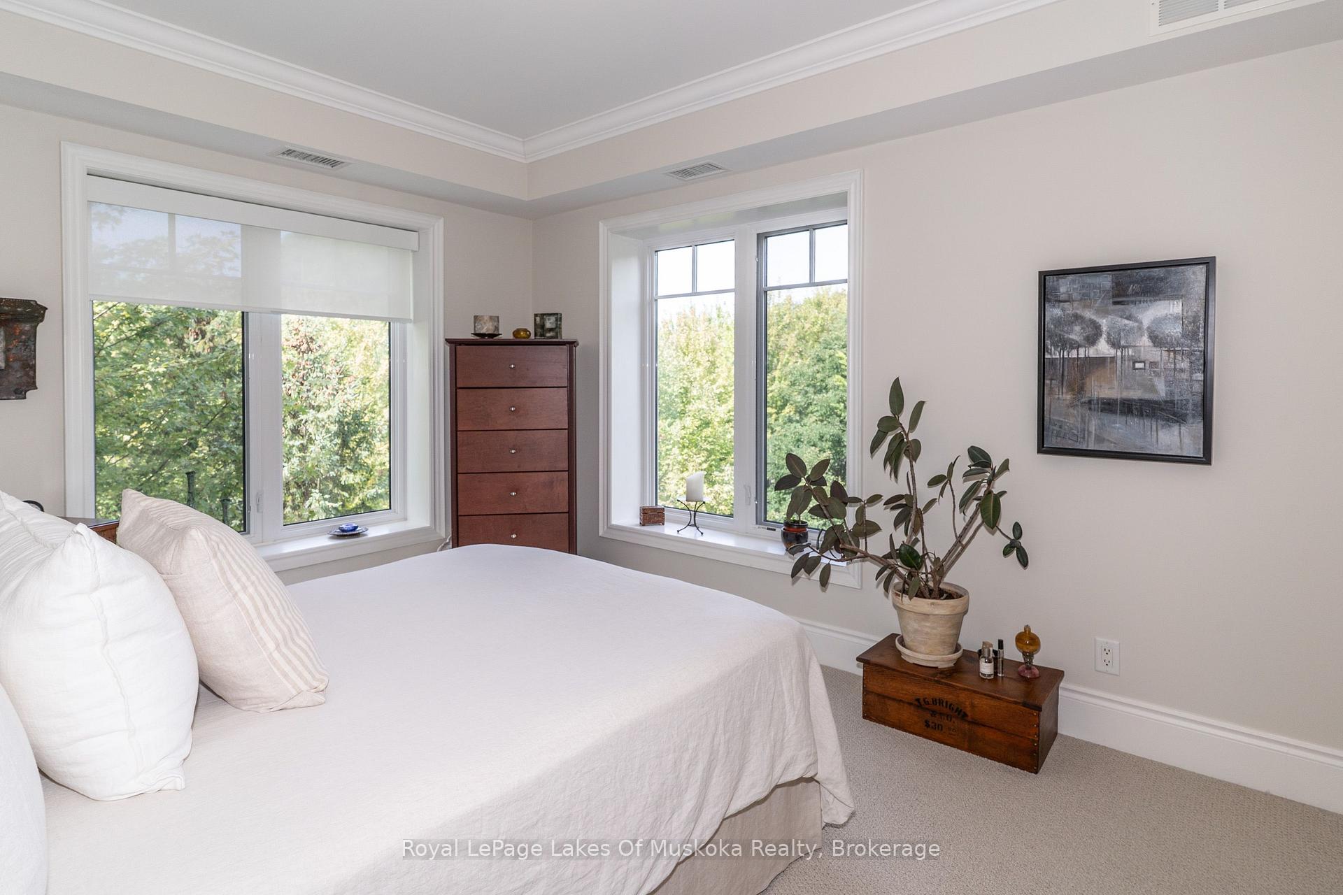 Bedroom with a white bed, tall dresser, and large windows overlooking trees.