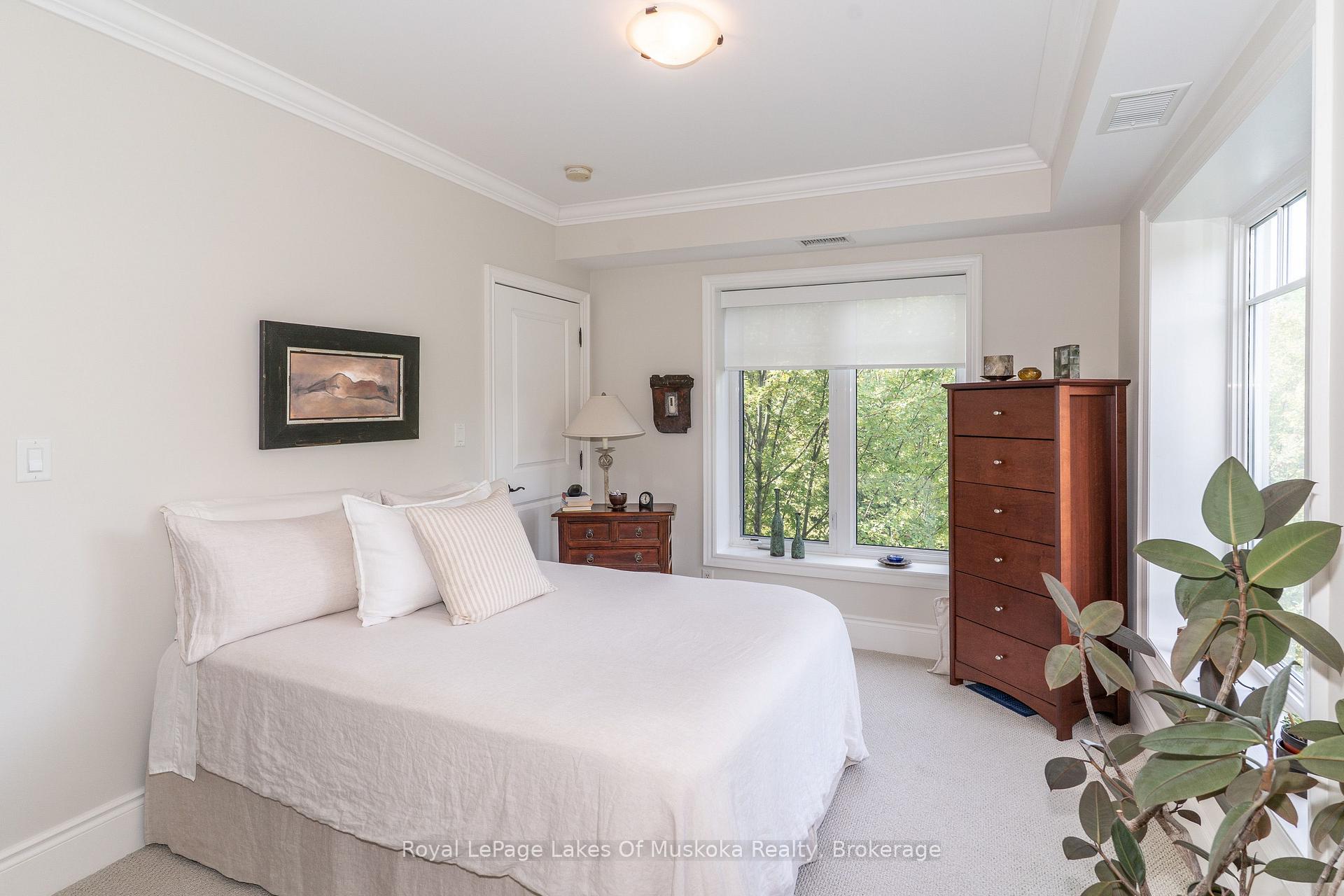 Bedroom with a white bed, dark wood furniture, and leafy views through two windows.