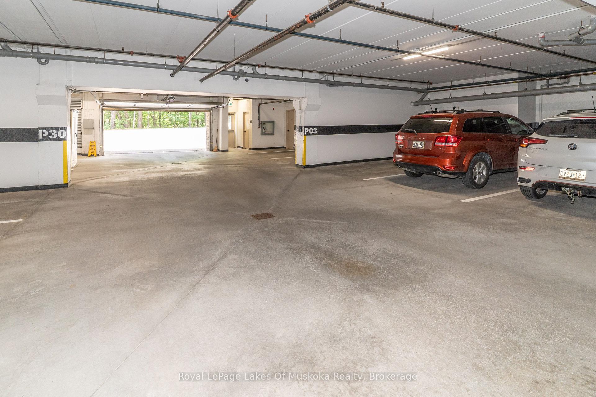 Condominium parking garage with open concrete spaces, exposed pipes, and two vehicles near the entrance.