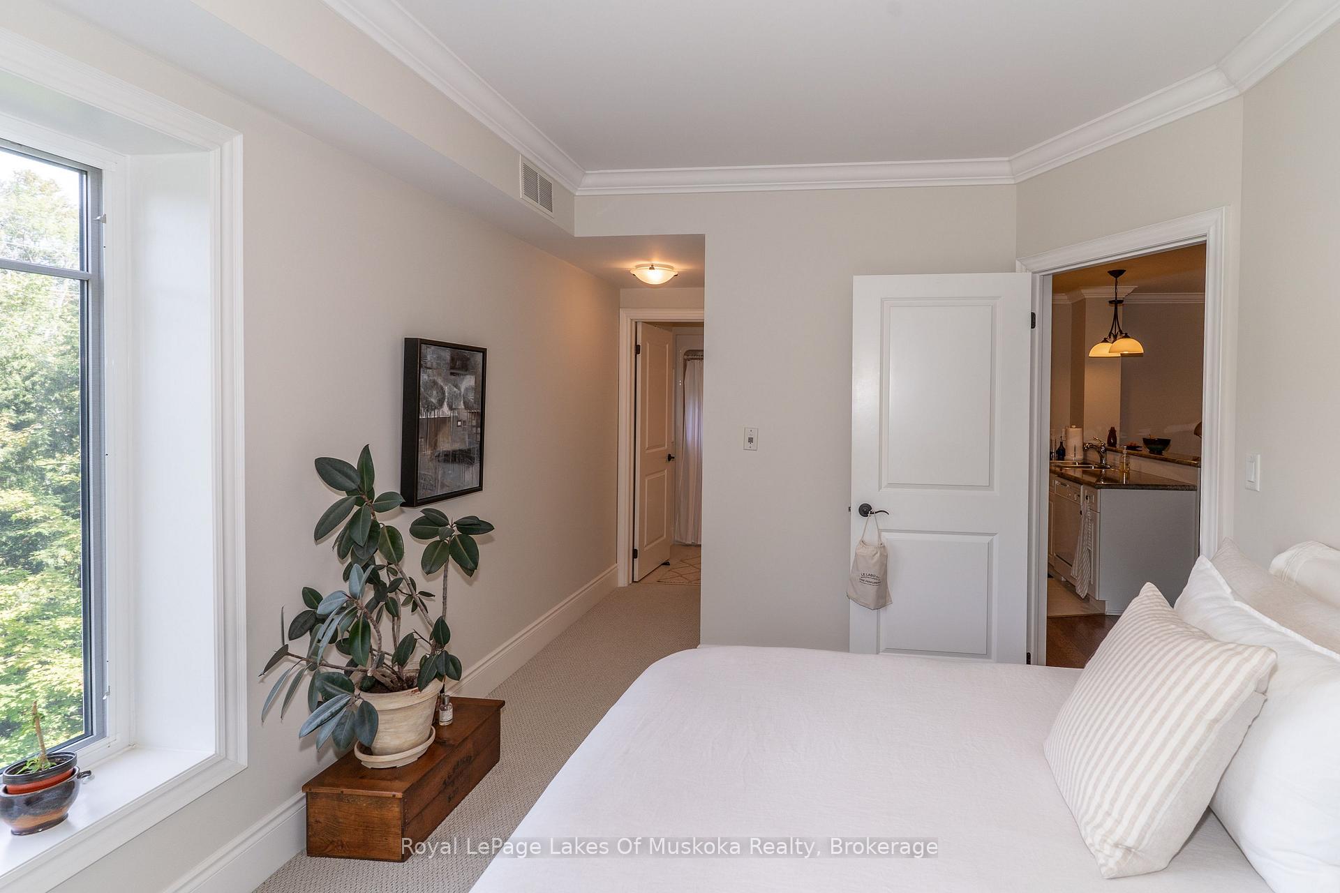 Bedroom with a white bed, large leafy-view window, and potted plant beside an open kitchen doorway.