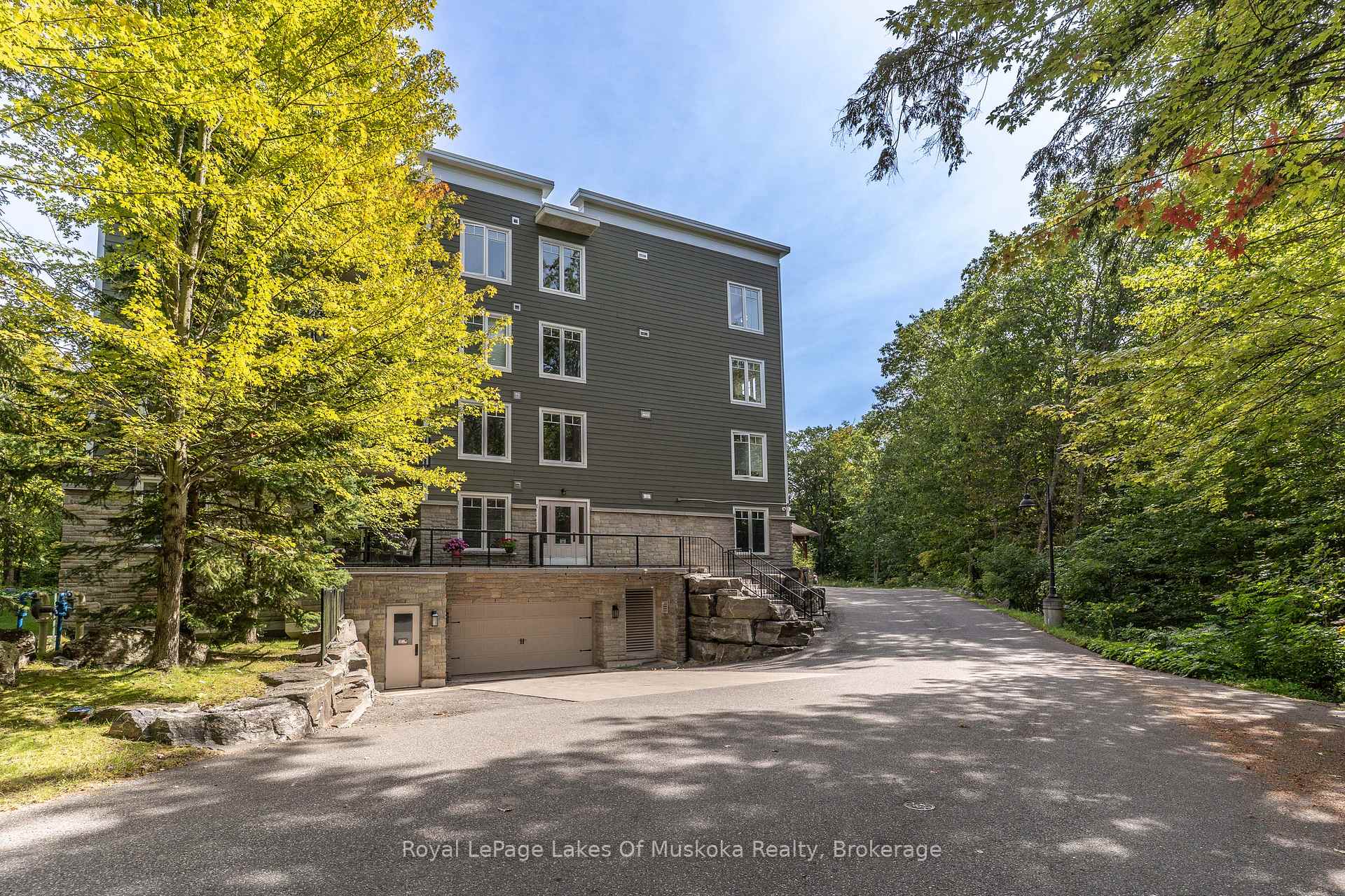 Multi-story building with dark siding, stone base, and garage beside a tree-lined driveway.