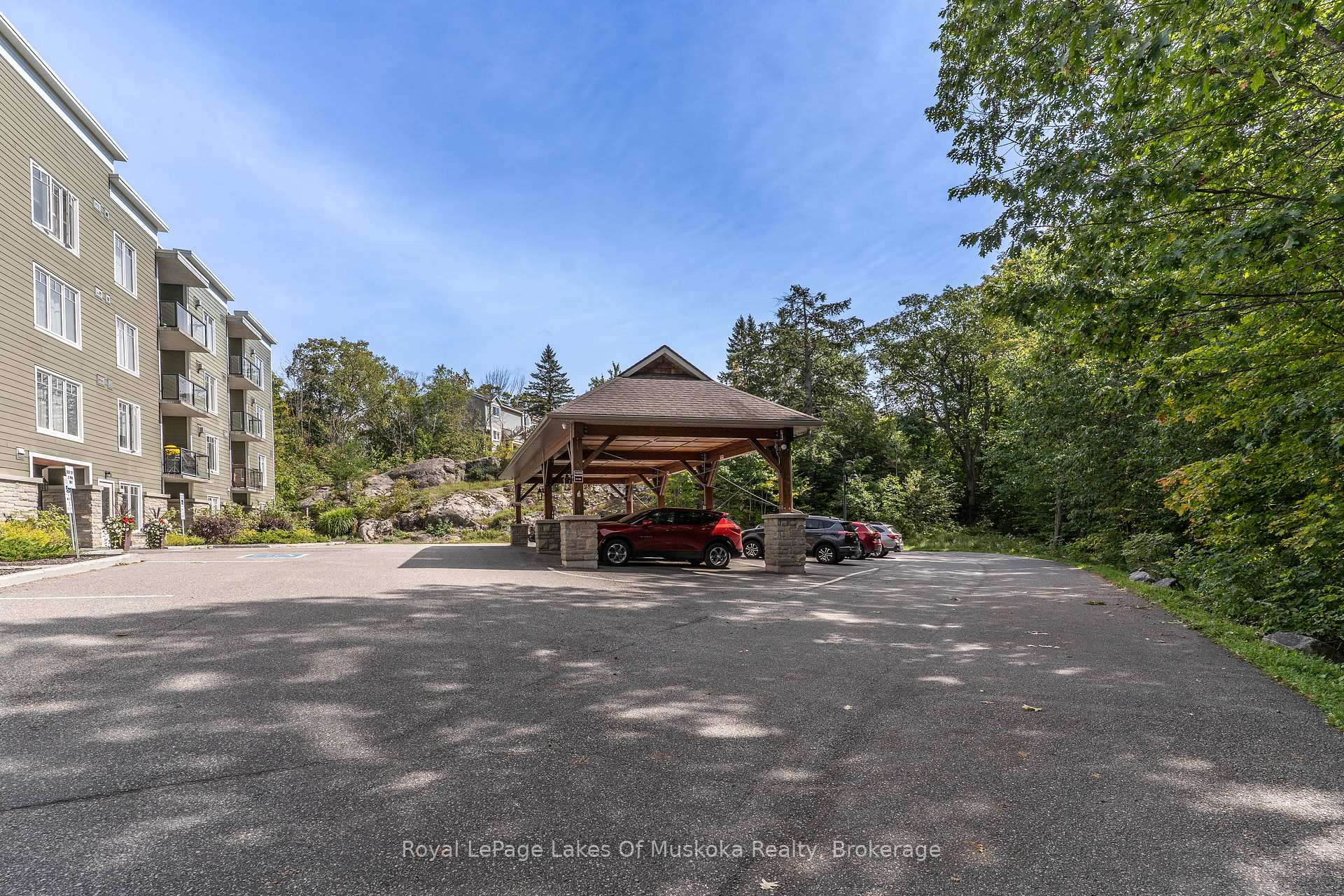 Condominium building with balconies beside a covered drop-off canopy and wooded parking area.