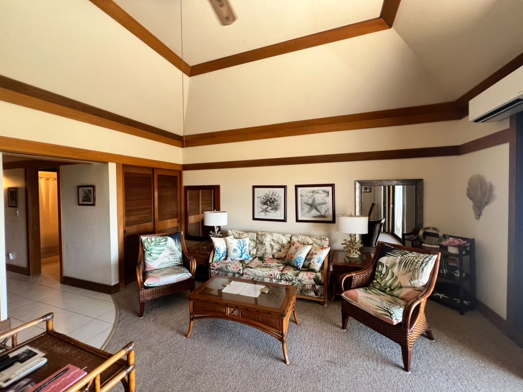 Living room with vaulted ceiling, dark wood trim, wicker furniture, and tropical-print cushions.