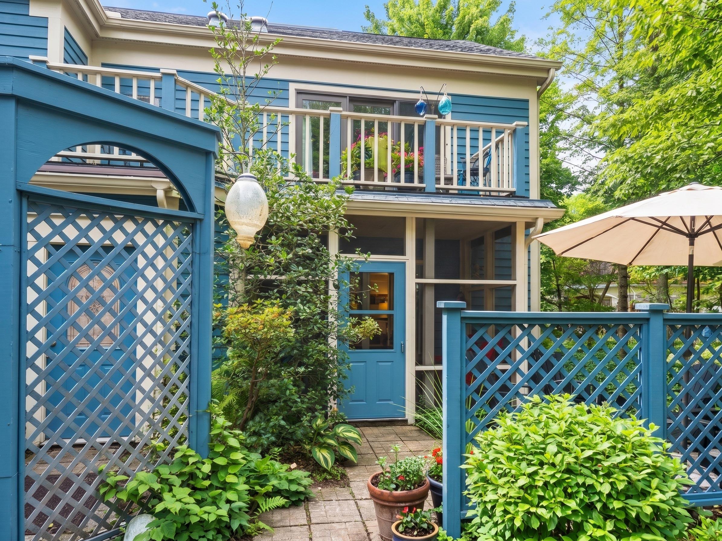 Blue-sided home with an upper balcony, screened porch, and lattice fencing around a leafy garden.