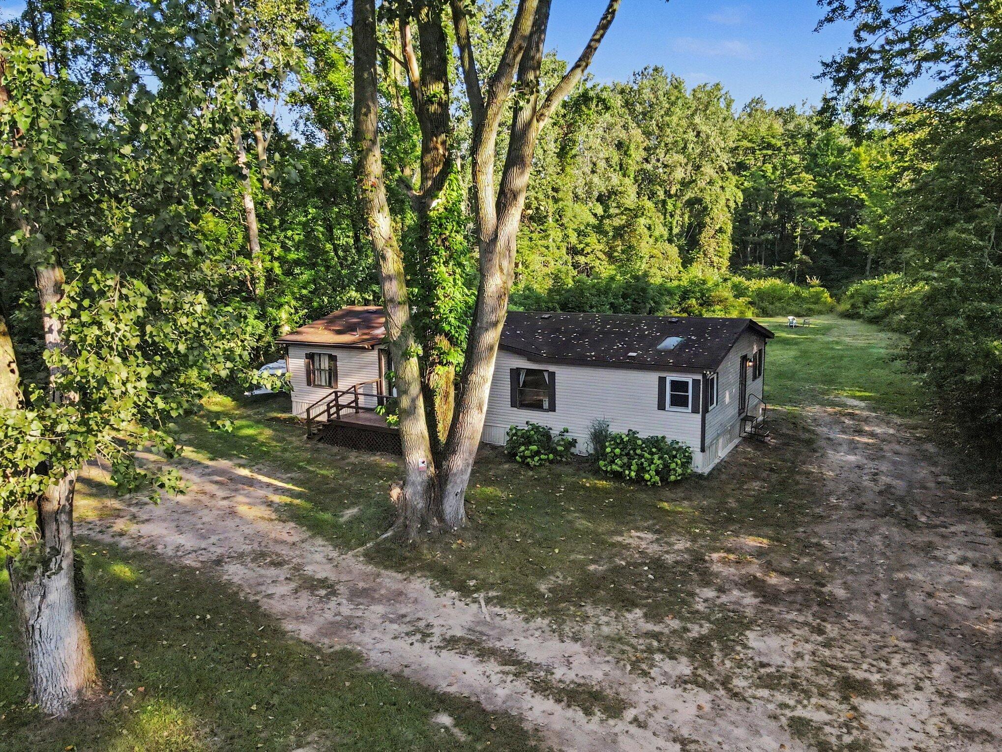 Aerial view of a light-sided manufactured home surrounded by mature trees, with a sandy driveway and open lawn.