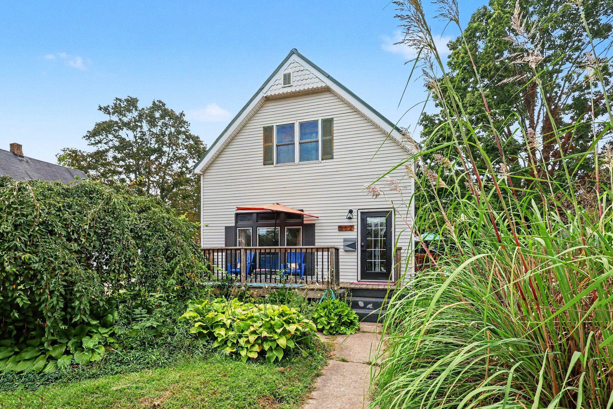 Two-story gabled house with a small deck, blue seating, and a garden path through lush greenery.