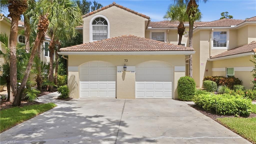 Two-story beige home with a tile roof, double garage, wide driveway, and tropical landscaping.