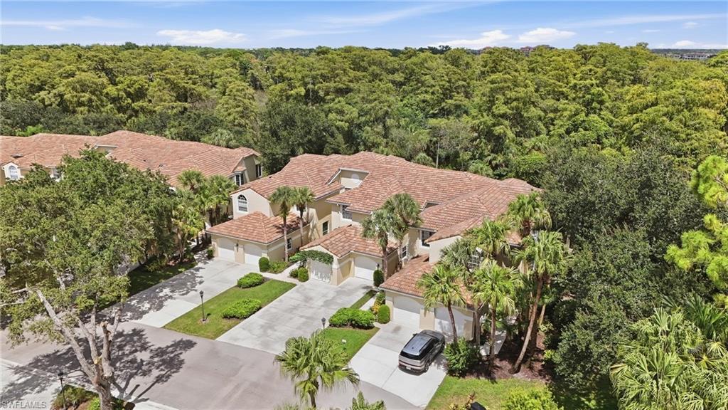 Aerial view of a tile-roofed home with multiple garages, palms, and dense trees behind it.