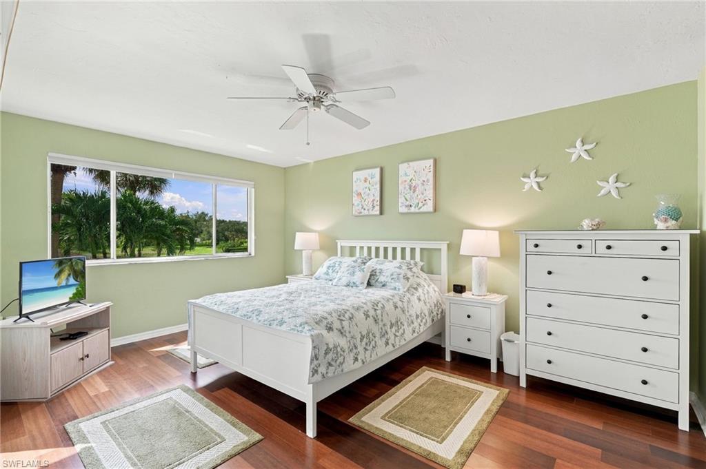Furnished bedroom with floral bedding, pale green walls, and a wide window overlooking palm trees.