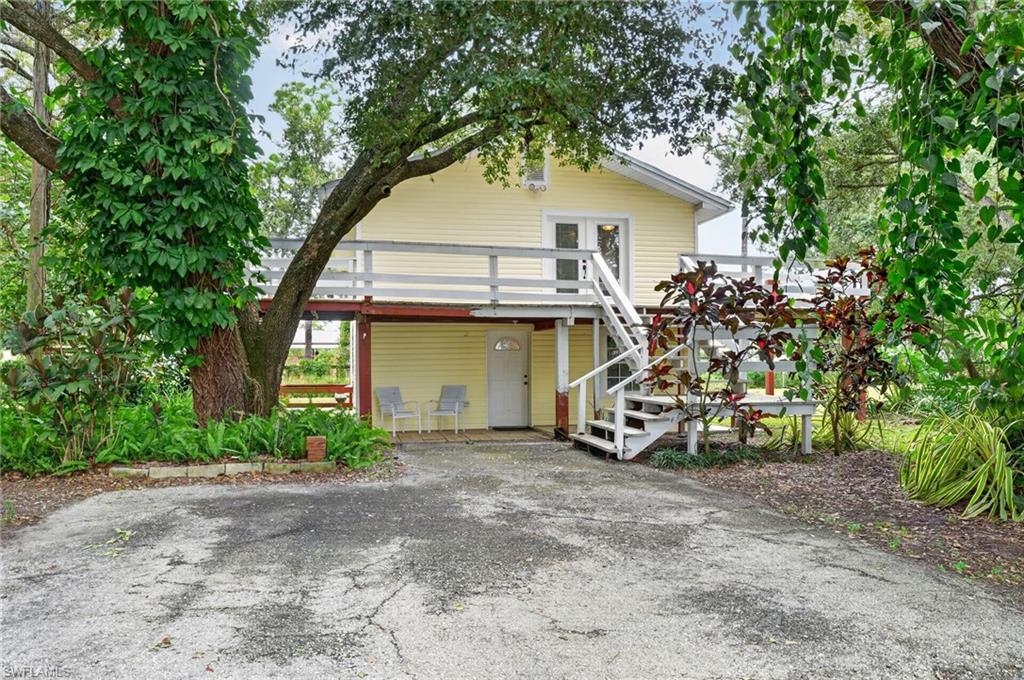 Yellow raised house with a white-railed deck and staircase, surrounded by mature trees and landscaping.