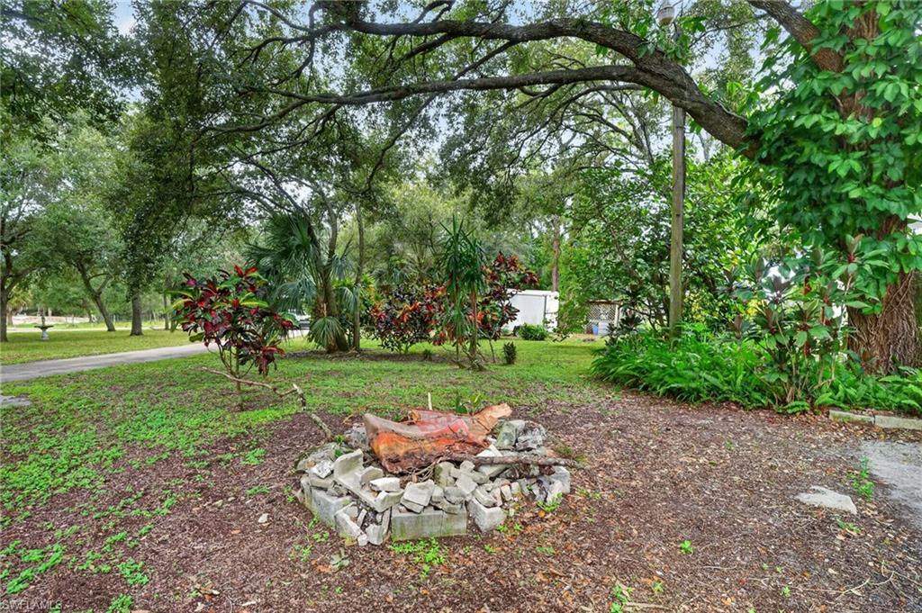 Shaded yard with a stone fire pit, mature trees, palms, and colorful shrubs.