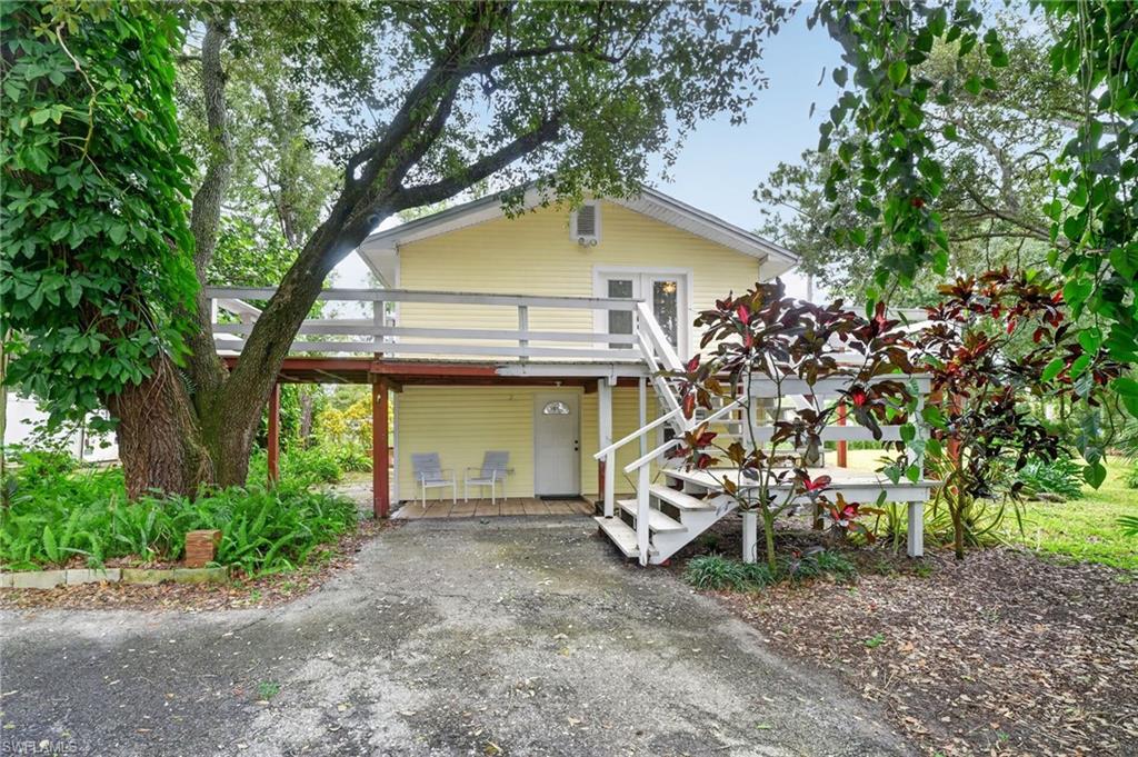 Pale yellow home with a white-railed deck, exterior stairs, and a shaded patio beneath mature trees.