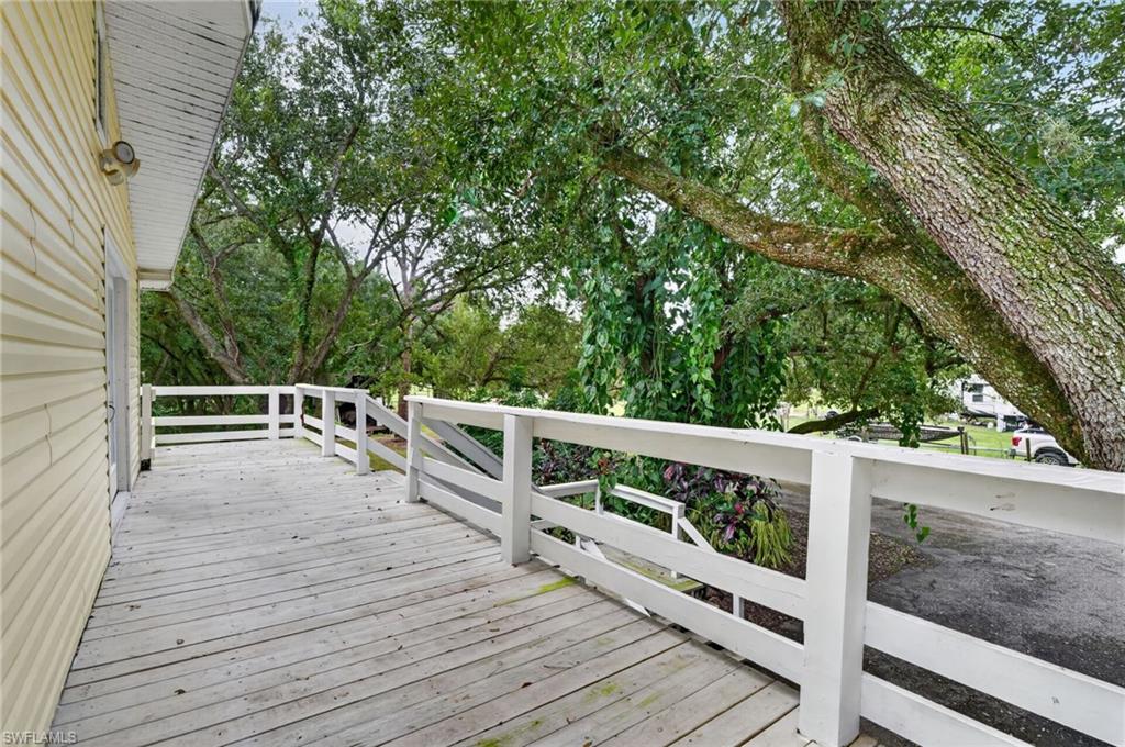 Wooden deck with white railings beside a pale yellow home, shaded by large leafy trees.