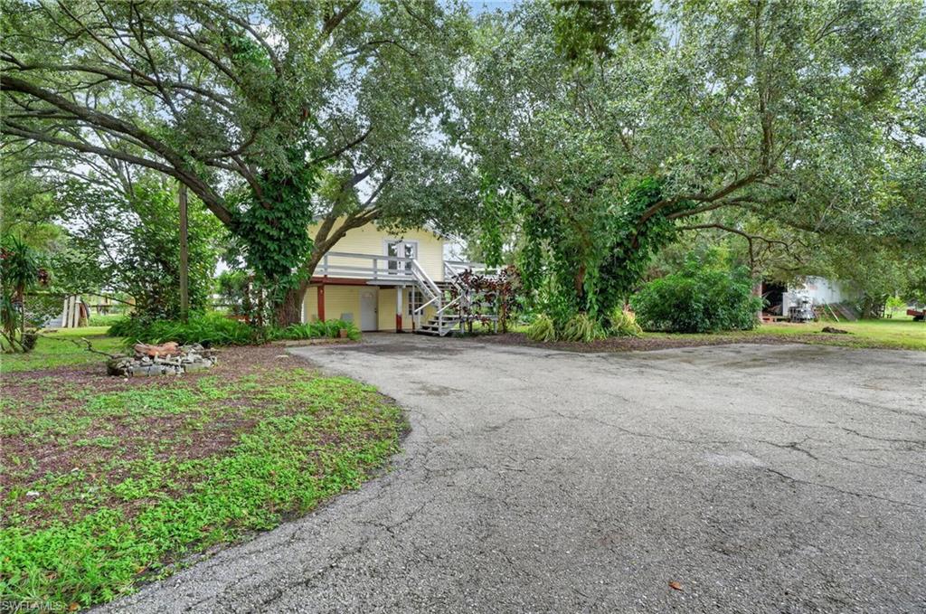 Light-yellow elevated home beneath mature trees, with an outdoor staircase and broad paved driveway.