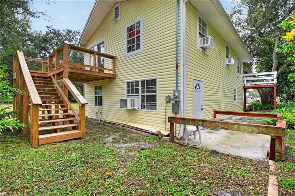 Pale yellow two-story house with wooden stairs and deck beside a concrete patio.
