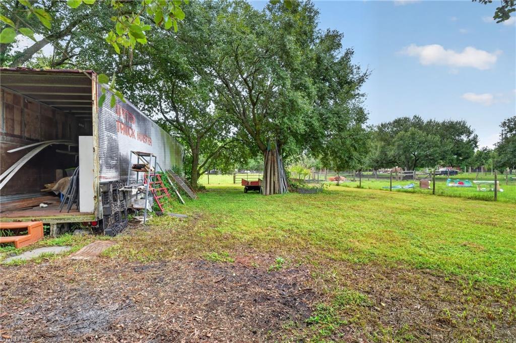 Grassy rural yard with mature trees, fenced grounds, and an open-sided storage trailer.