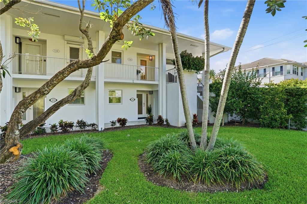 Two-story white residential building with balconies, framed by palm trees and a landscaped lawn.