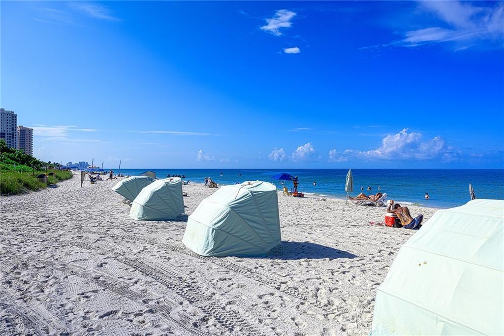 Sandy beach with covered cabanas, beachgoers, blue ocean, and distant high-rise buildings.