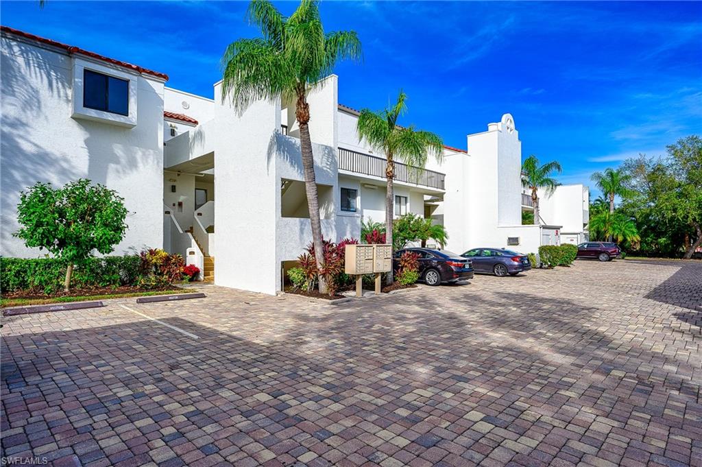 White stucco condominium building with palm trees, balconies, and a broad paver parking area.