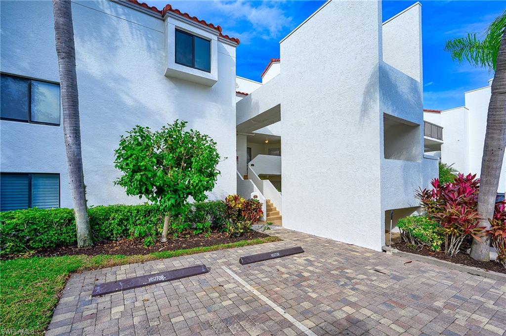 White stucco condominium entrance with red tile roof accents, tropical landscaping, and visitor parking.