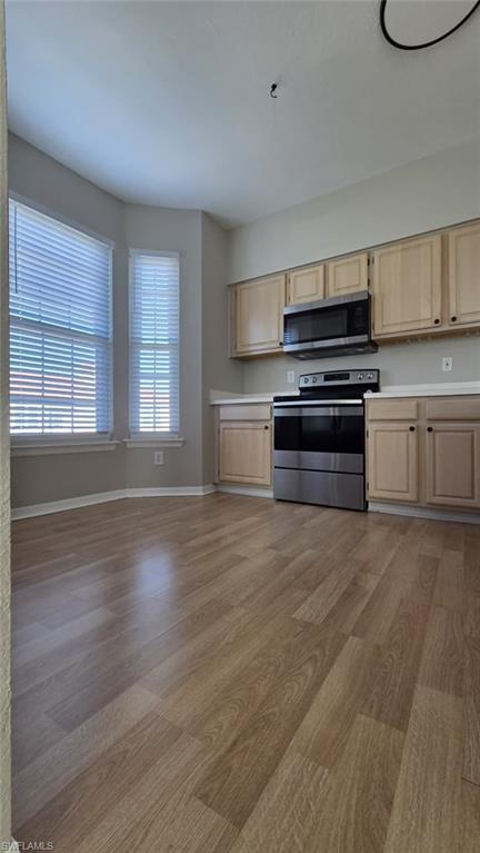 Kitchen with light wood cabinets, stainless steel appliances, wood-look floors, and two tall windows with blinds.
