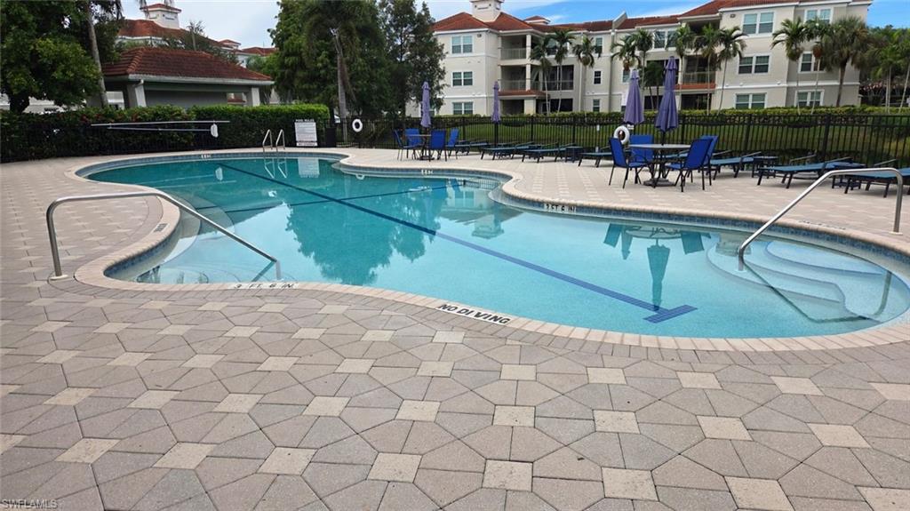 Community pool with a curved shallow-entry area, paver deck, blue lounge chairs, and residential buildings.