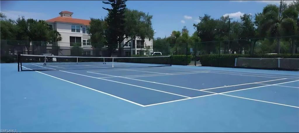 Outdoor blue tennis court with a net, hedges, and a terracotta-roofed building beyond.