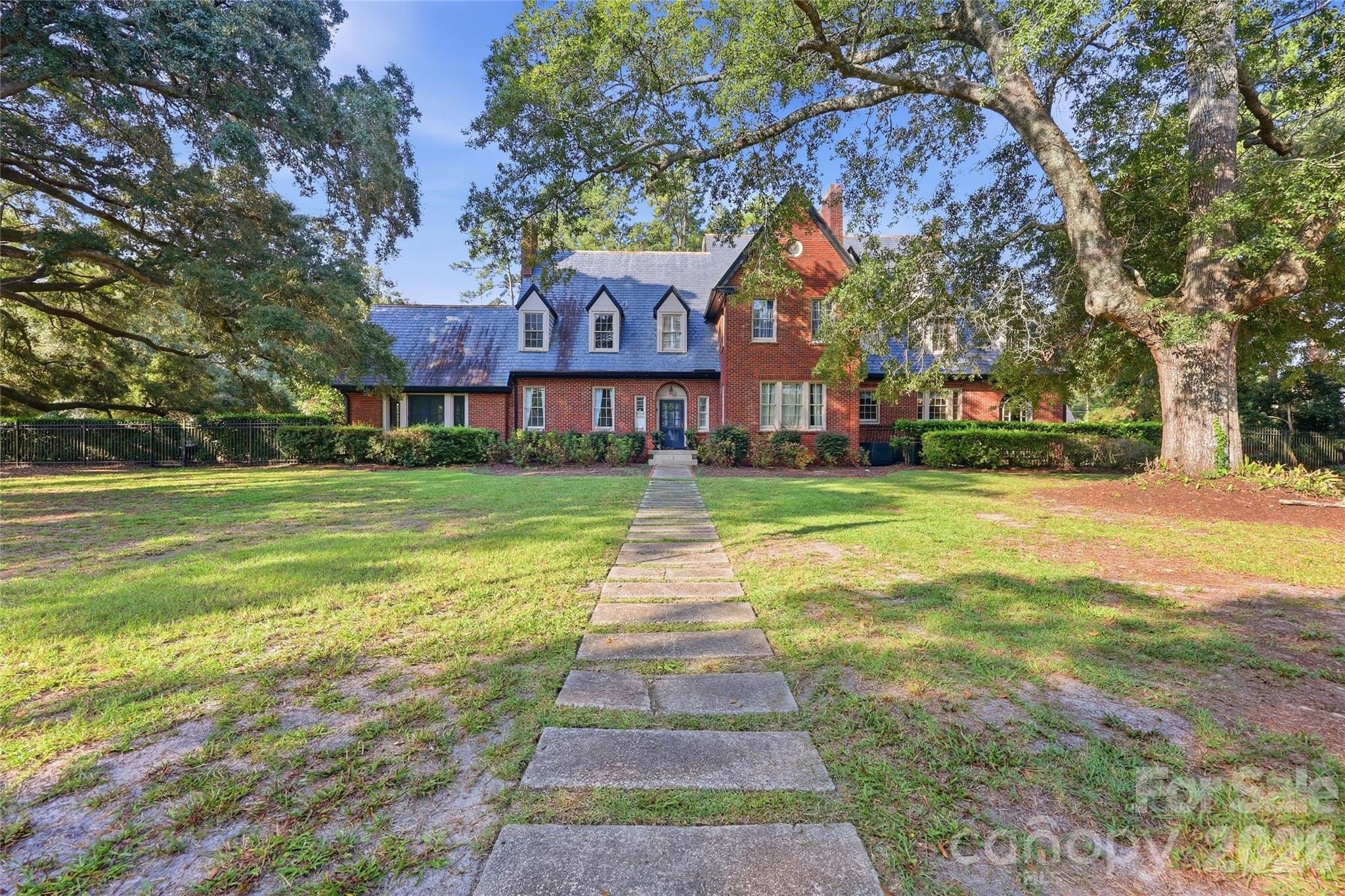 Red-brick home with dormer windows, framed by mature oaks and a stone front walkway.