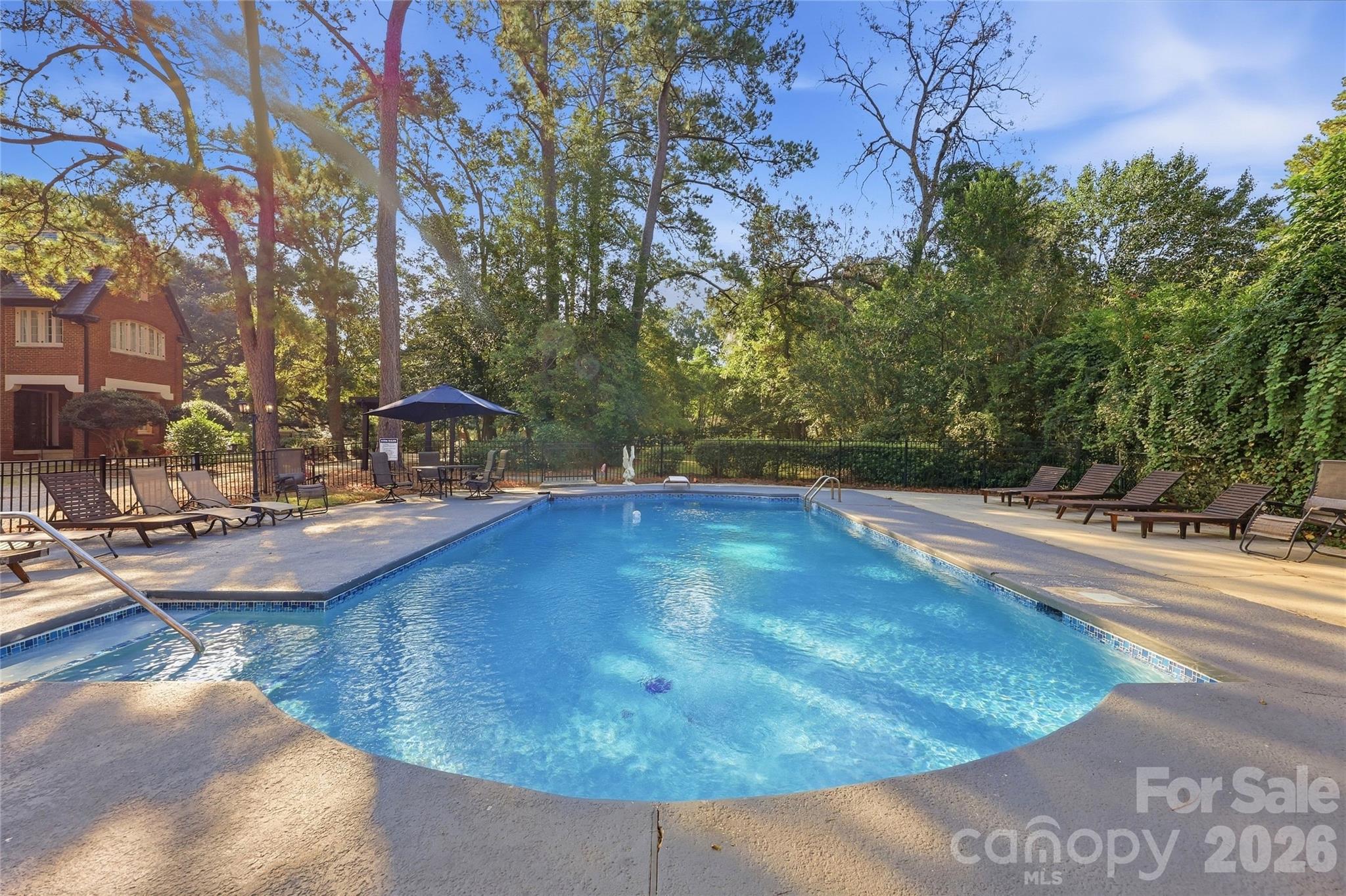 Outdoor pool with a curved deck, lounge chairs, umbrella-shaded seating, and mature trees.