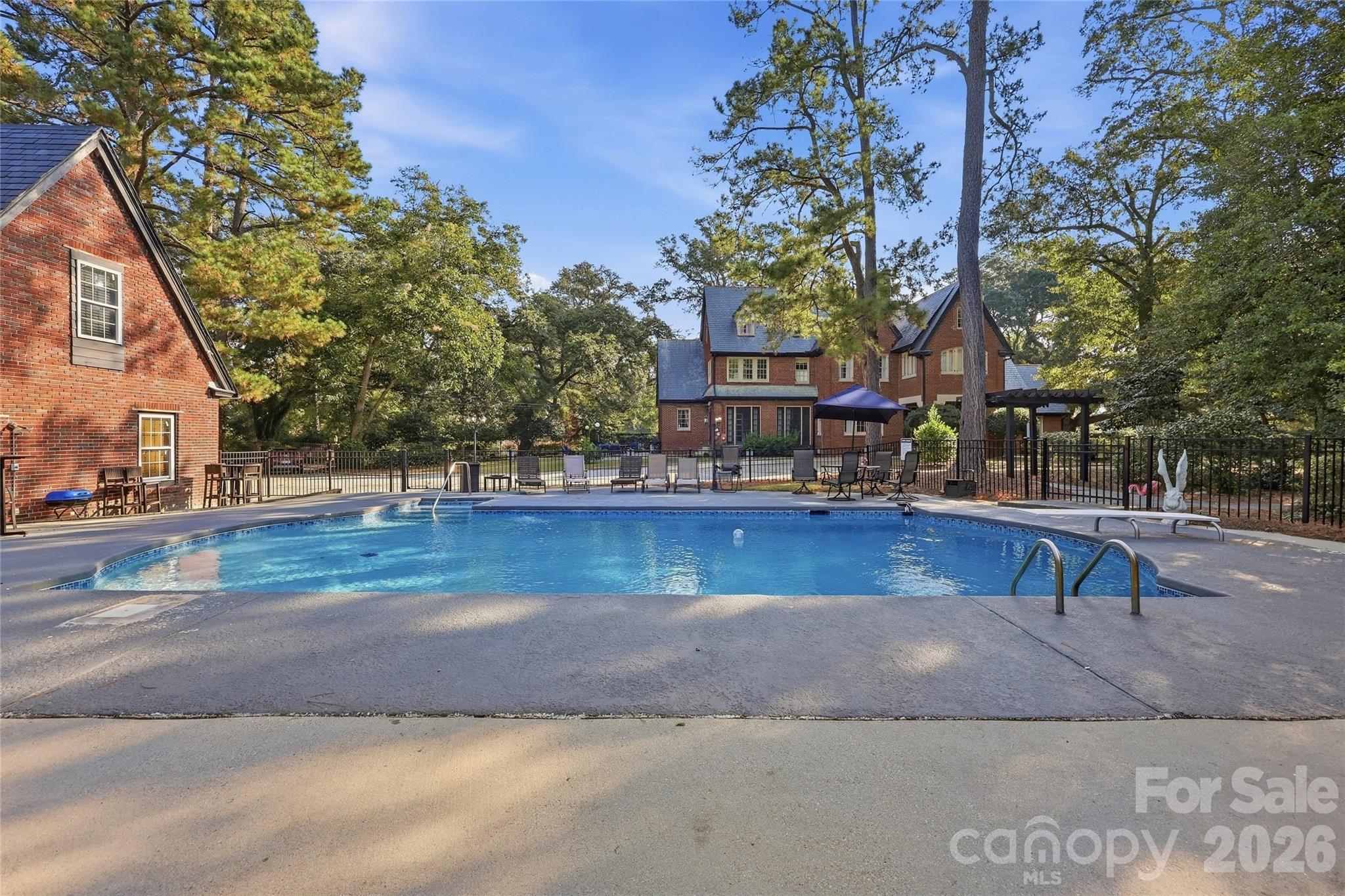 Outdoor pool beside brick homes, with patio seating, mature trees, and a fenced concrete deck.