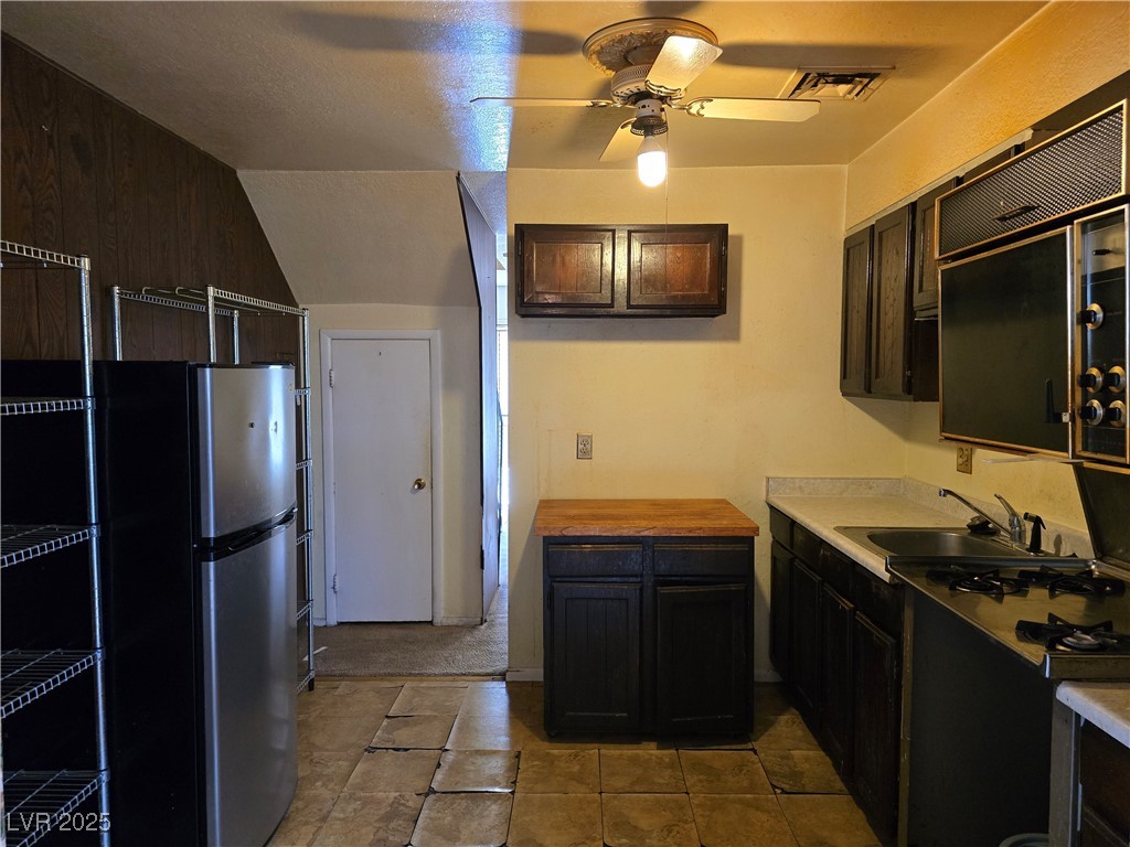 Narrow kitchen with dark cabinets, stainless steel refrigerator, gas cooktop, and a small wood-top island.