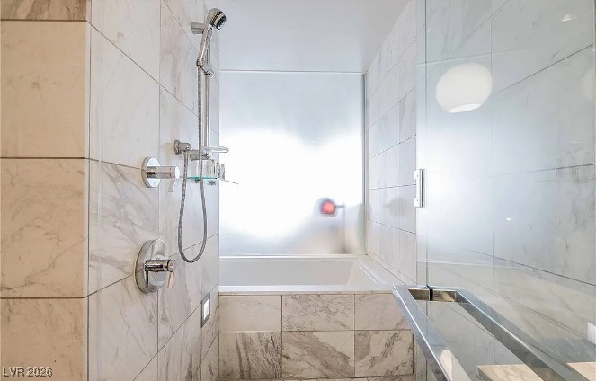 Marble-tiled bathtub and shower with a glass enclosure and handheld fixture.