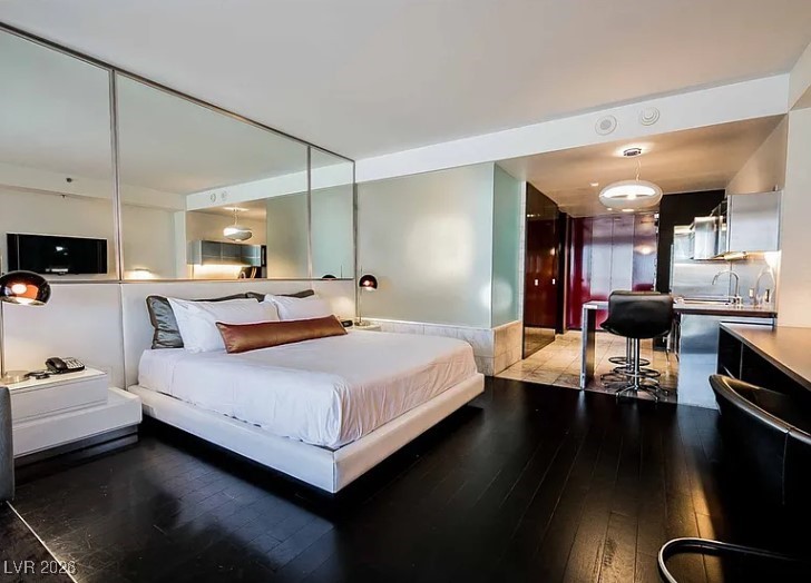Studio bedroom with a white bed, mirrored wall, dark wood floors, and an adjoining kitchenette.