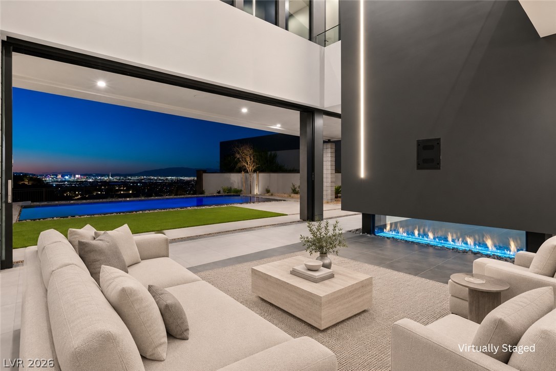 Living room with a linear fireplace and wide opening to a pool, lawn, and city skyline at dusk.