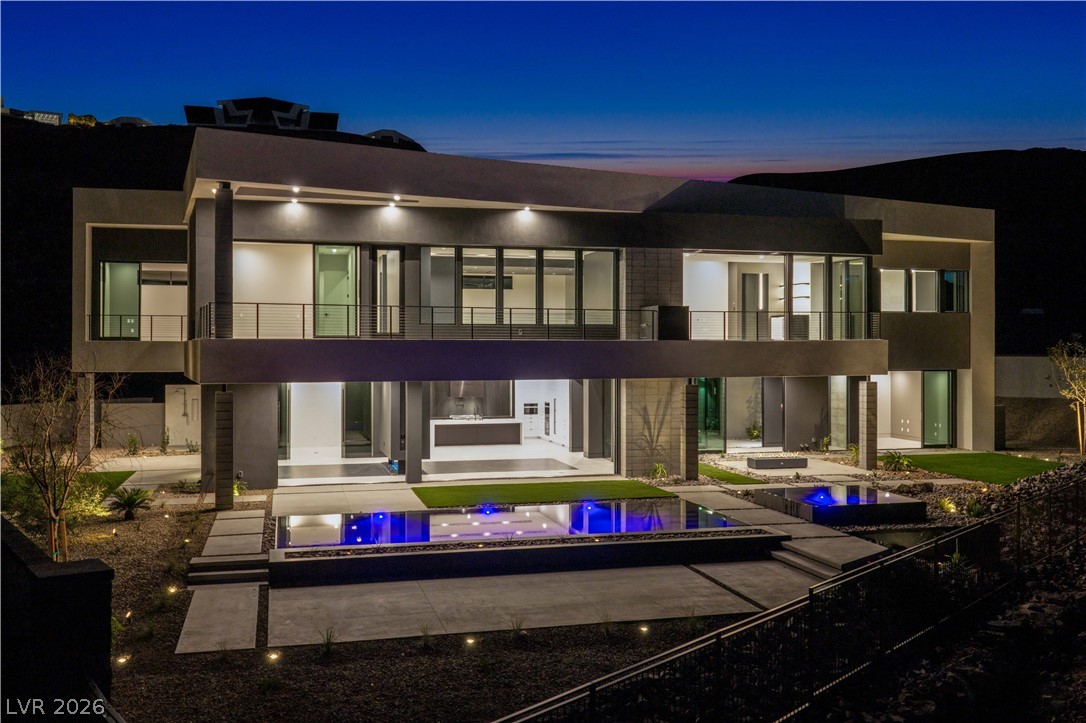 Modern two-story home with a glass-fronted balcony and blue-lit pool at dusk.