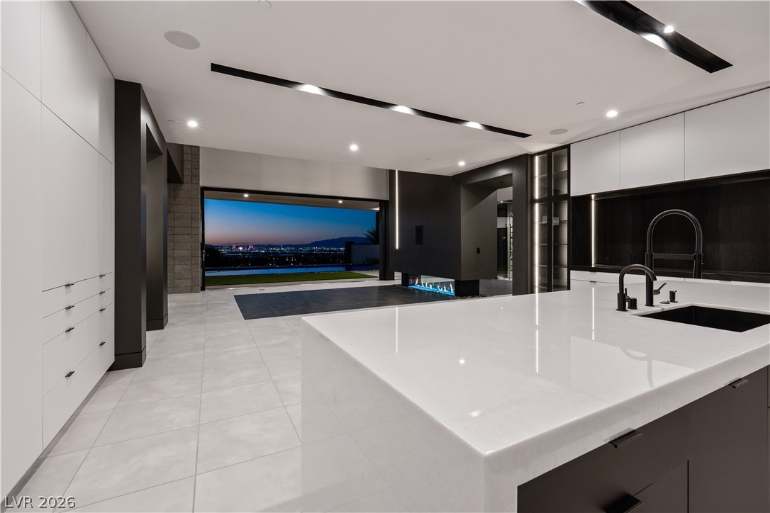 Modern kitchen with a large white island opening to a pool and city lights at dusk.