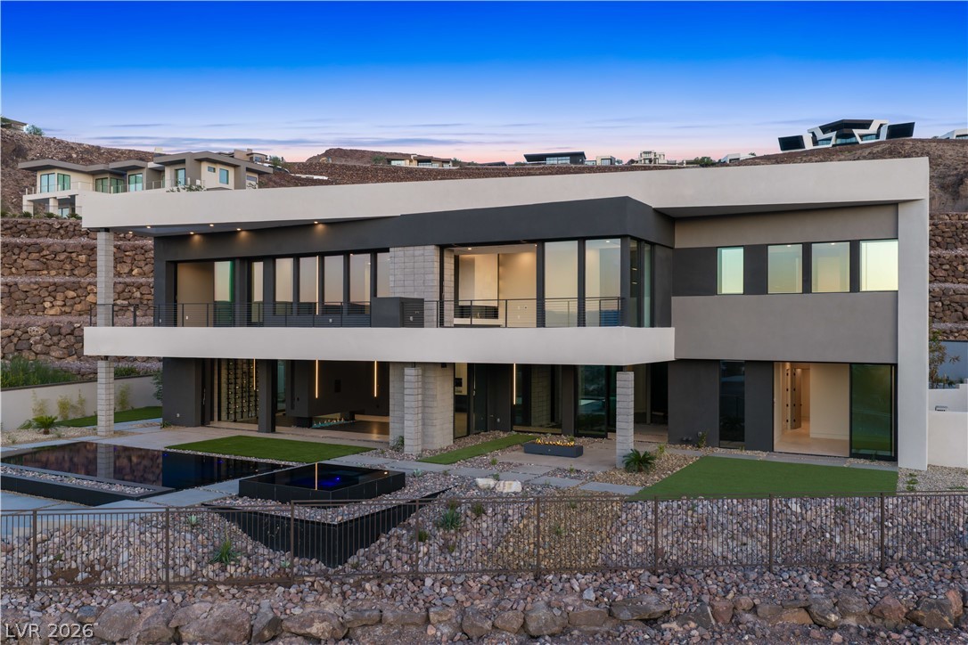 Modern two-story home with a broad balcony, swimming pool, and desert rock landscaping at dusk.