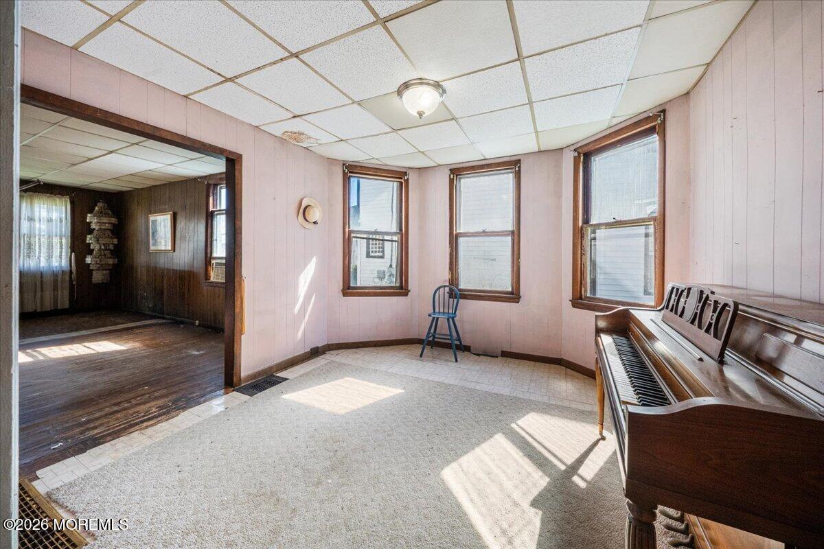 Interior room with pink paneled walls, three windows, and an upright piano beside a doorway to a dark wood-paneled room.