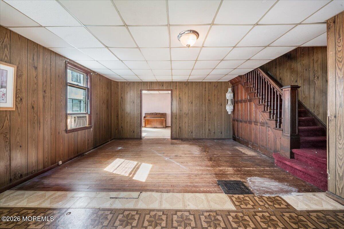 Vacant wood-paneled room with a red-carpeted staircase and a piano visible through the doorway.