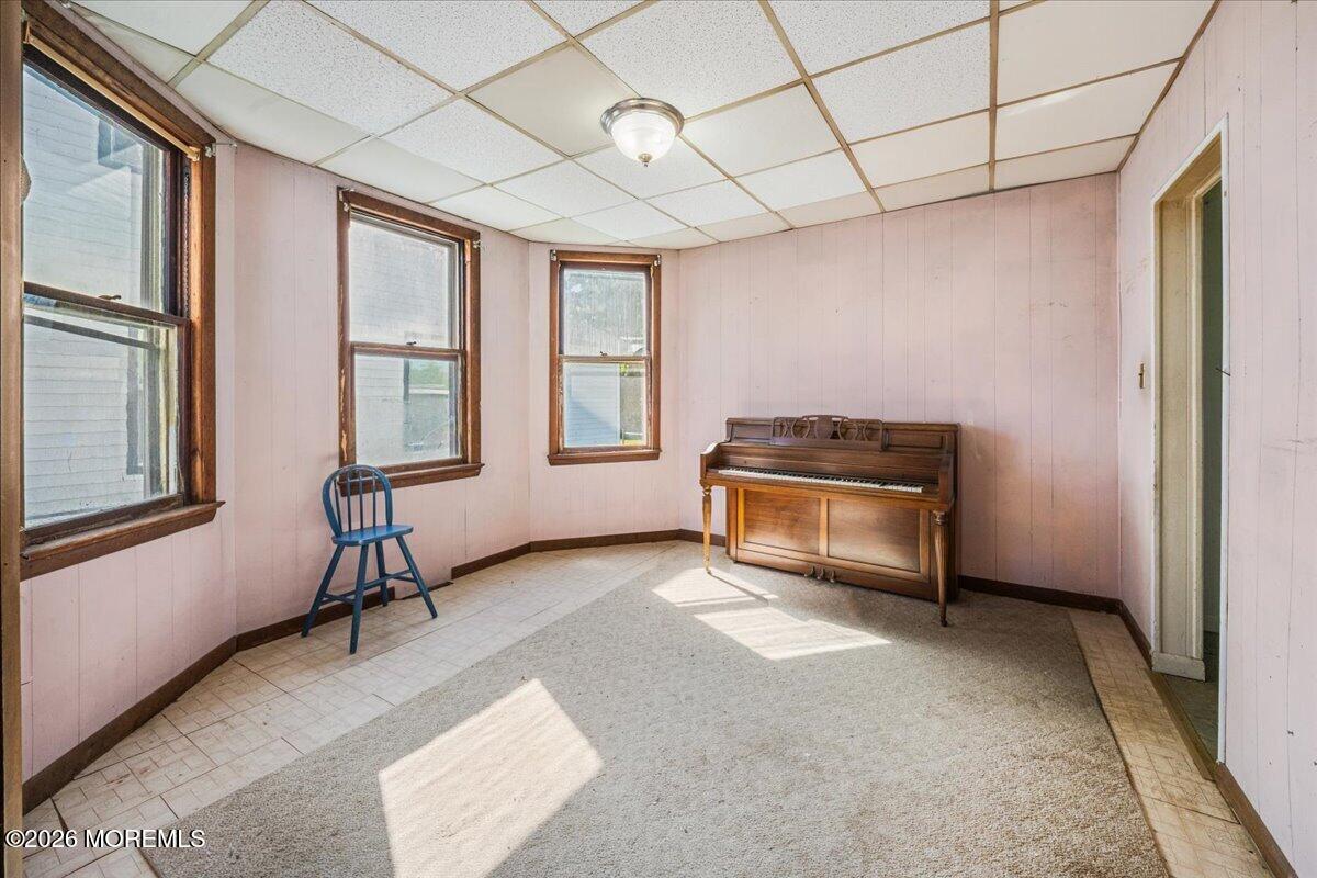 Room with a piano beneath three wood-trimmed windows and pale pink paneled walls.