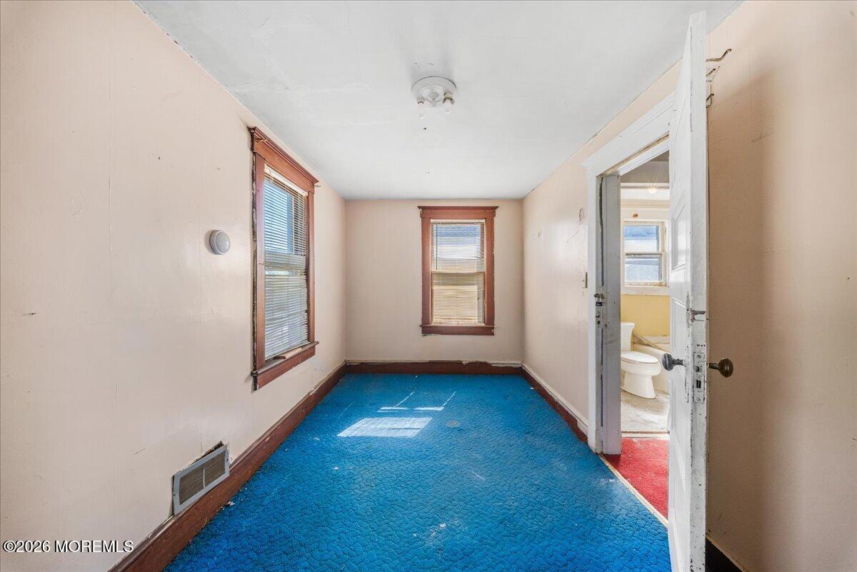 Empty narrow room with blue carpet and dark wood-trimmed windows, opening to a bathroom.