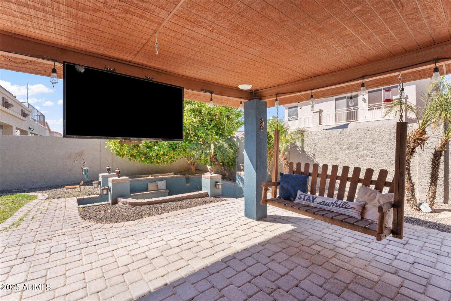 Covered patio with brick pavers, a hanging television, and a cushioned wooden swing bench.