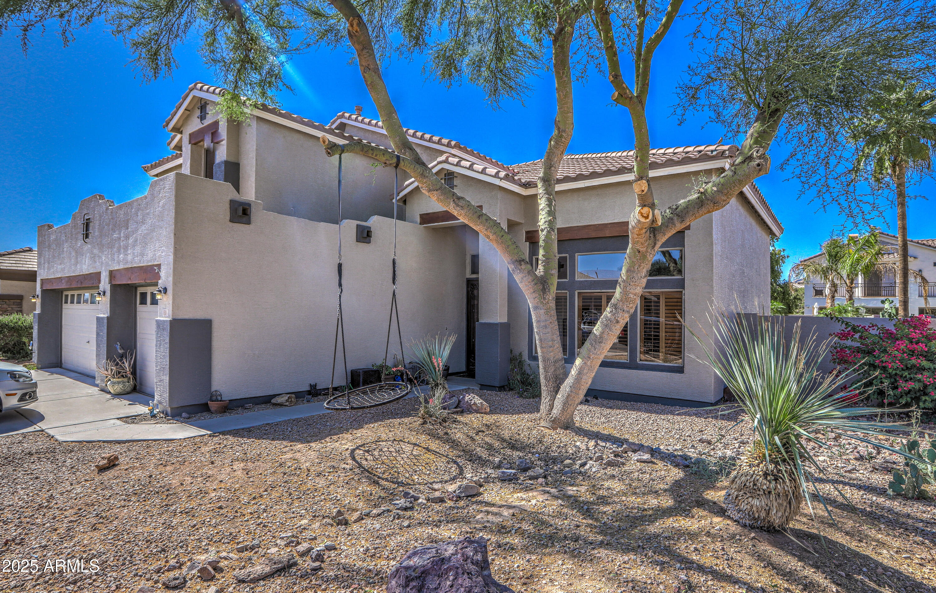 Two-story stucco home with a tiled roof, desert landscaping, and a hanging swing beneath a mature tree.