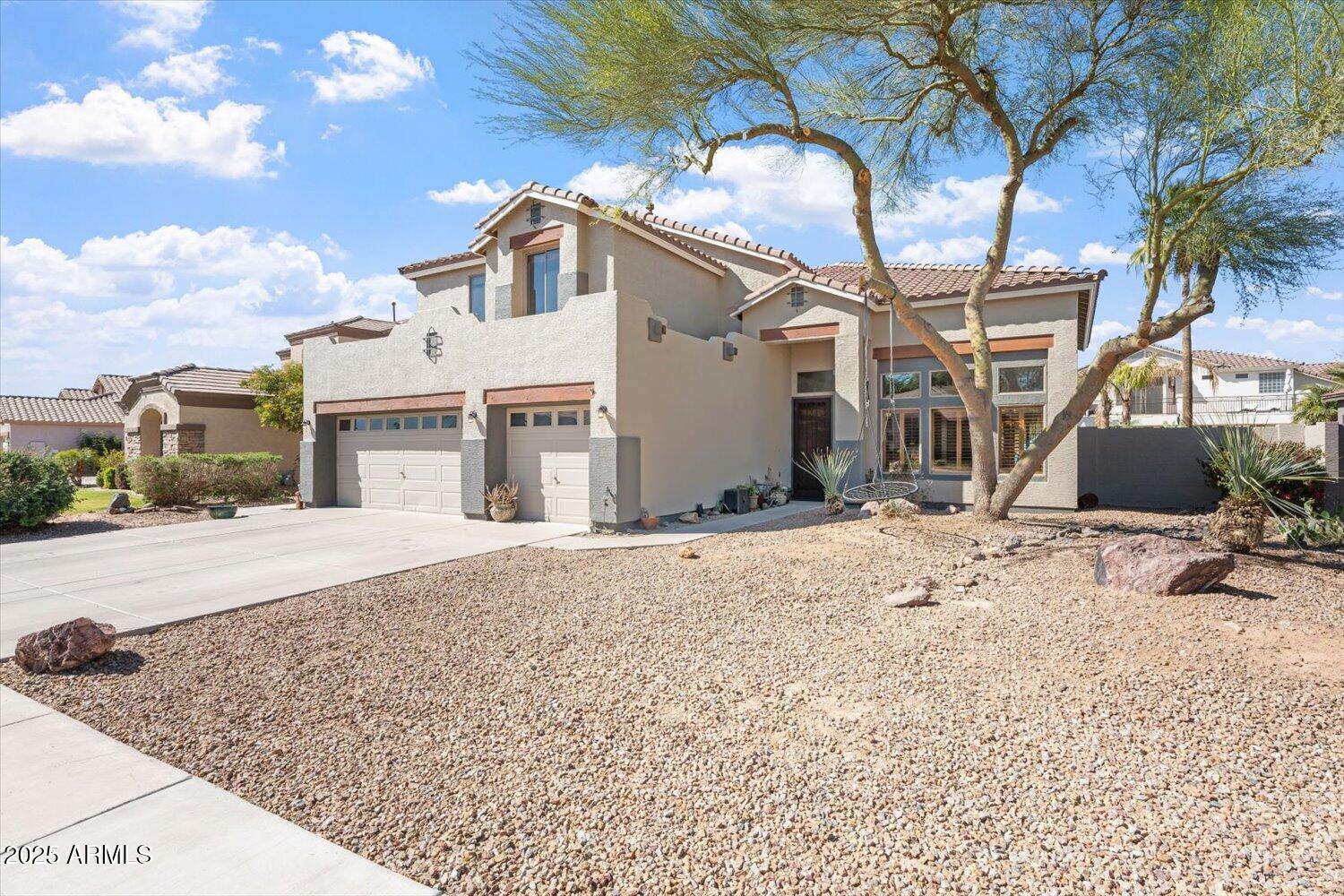 Two-story stucco home with a tile roof, two-car garage, and gravel landscaping beneath mature trees.