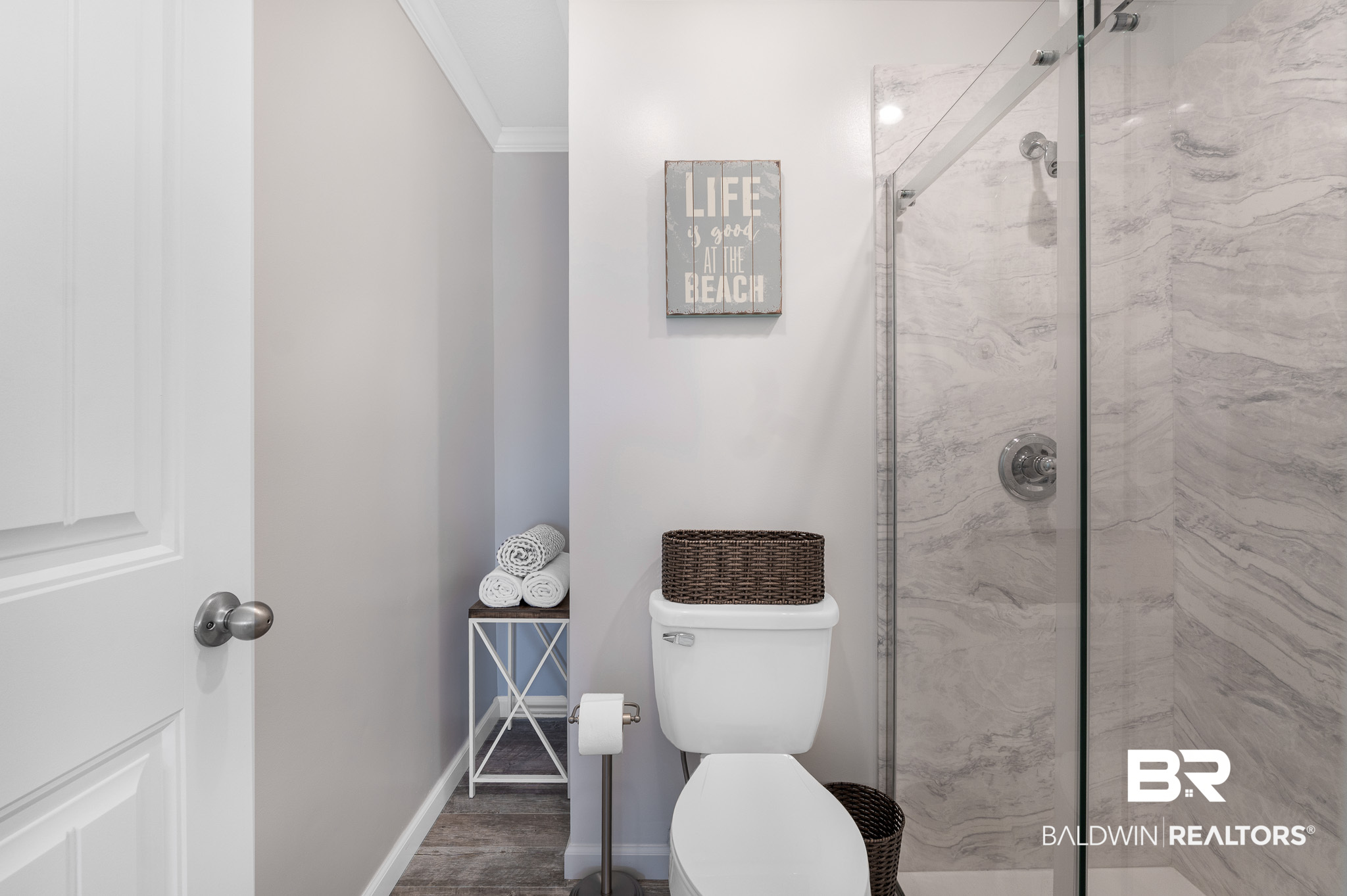 Bathroom with a glass marble shower, white toilet, rolled towels, and a beach-themed wall sign.